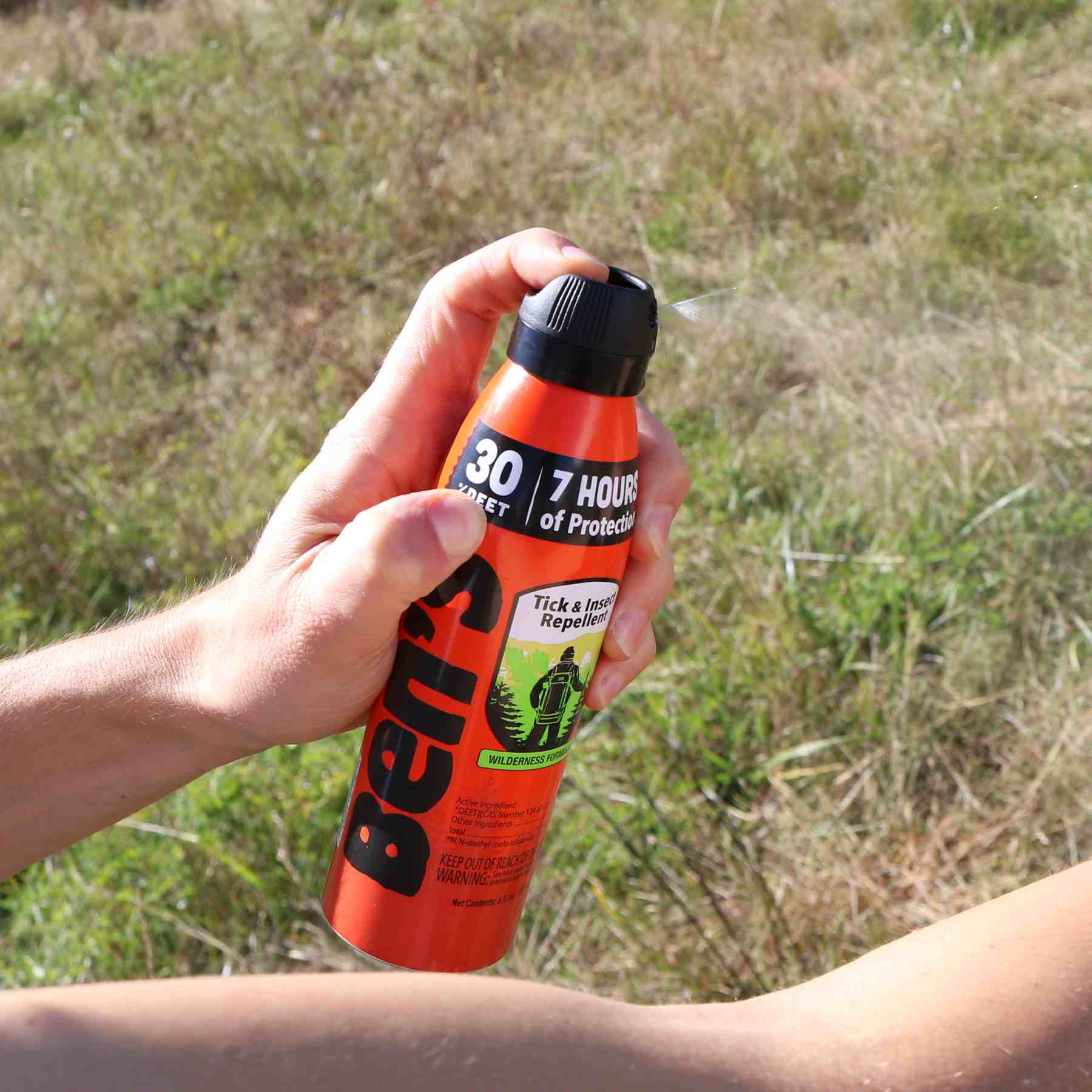 Person spraying Ben's 30 DEET 6 oz insect repellent on arm in front of grass