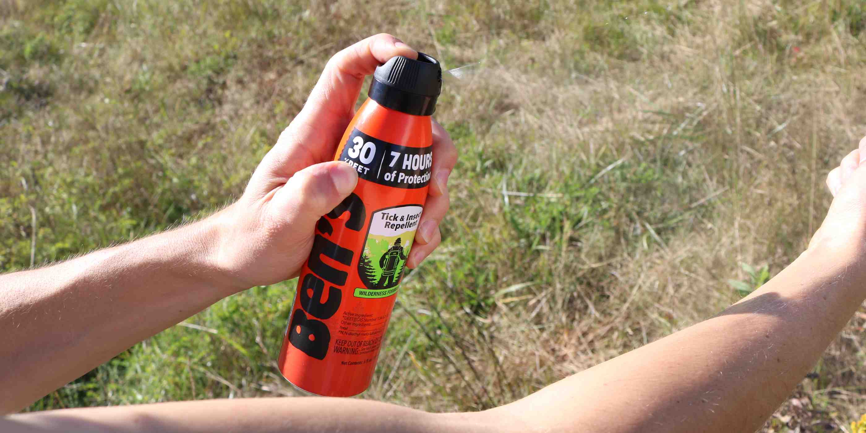 Person spraying Ben's 30 DEET 6 oz insect repellent on arm in front of grass