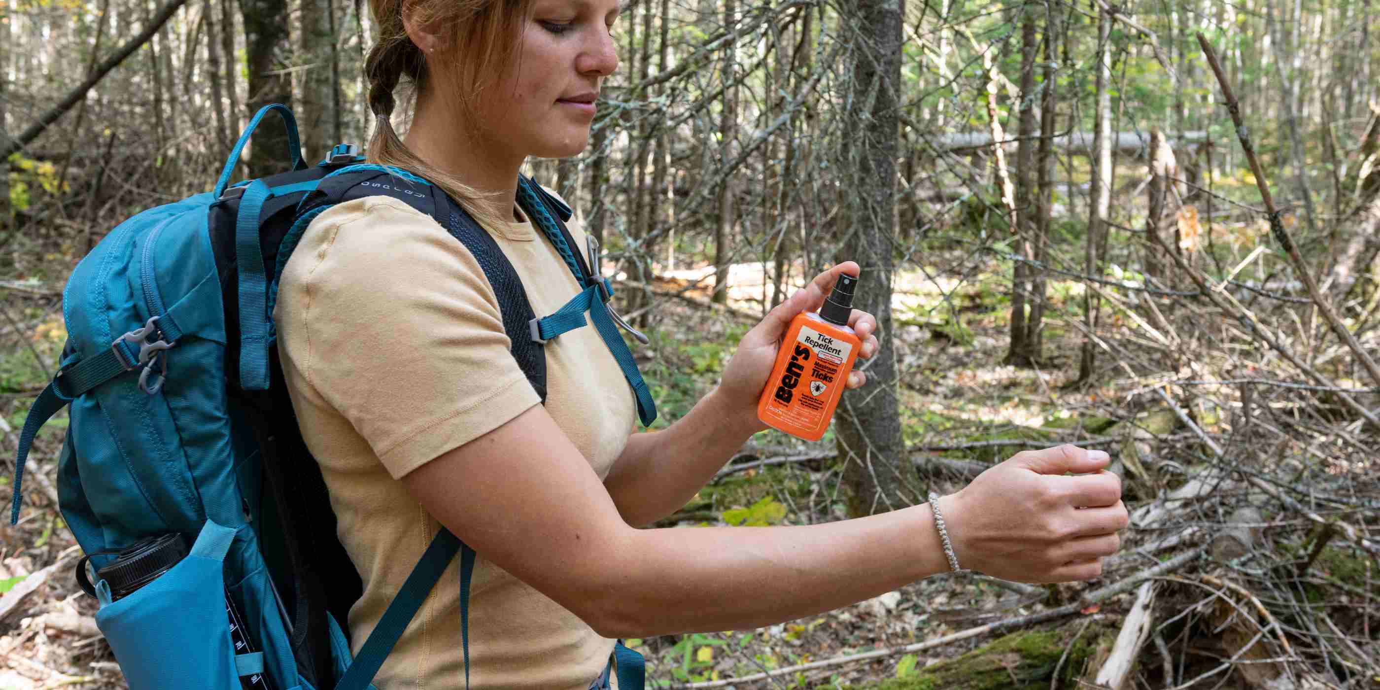 Woman using Ben's 3.4 oz Tick Repellent in woods with yellow shirt and blue backpack