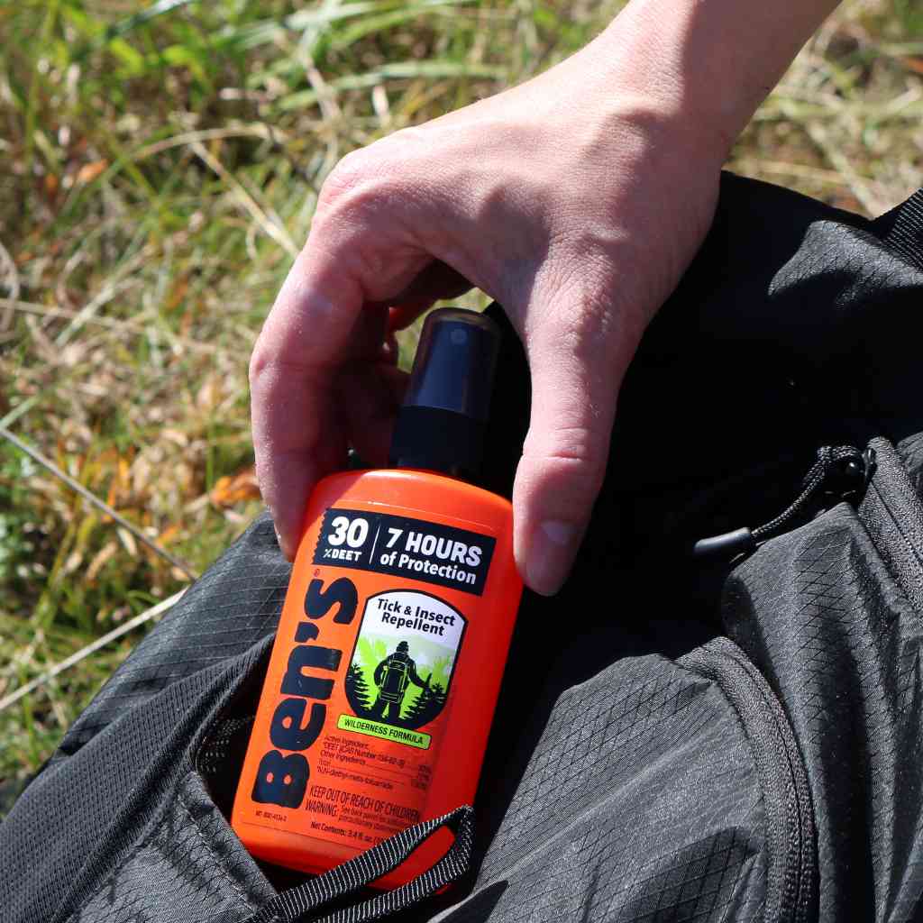 Ben's 30 Tick & Insect Repellent 3.4 oz. Pump Spray hand pulling from black backpack in front of grass
