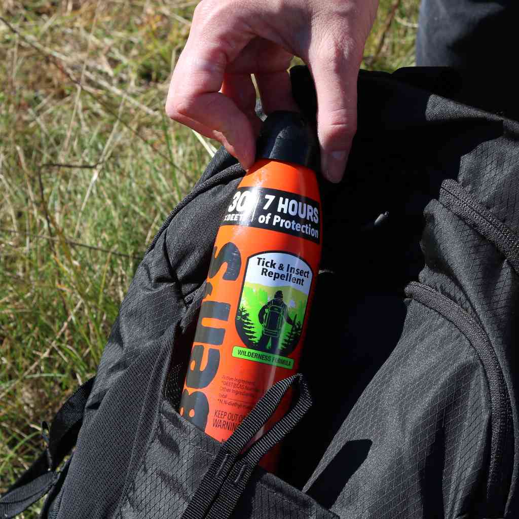 Ben's 30 Tick & Insect Repellent 6 oz. Eco-Spray hand pulling out of black backpack pocket in front of grass