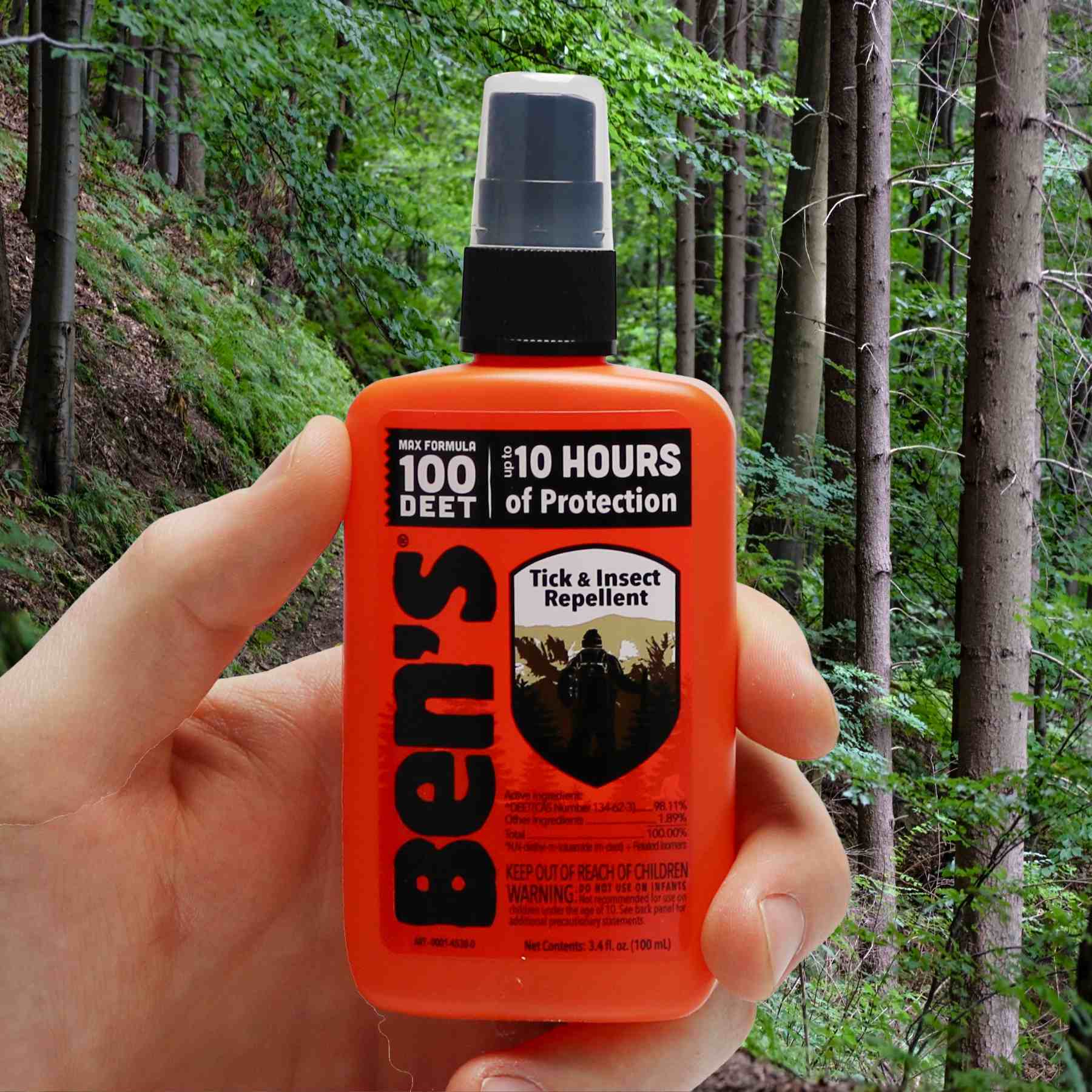 Hand holding Ben's 100 3.4 oz repellent in front of woods