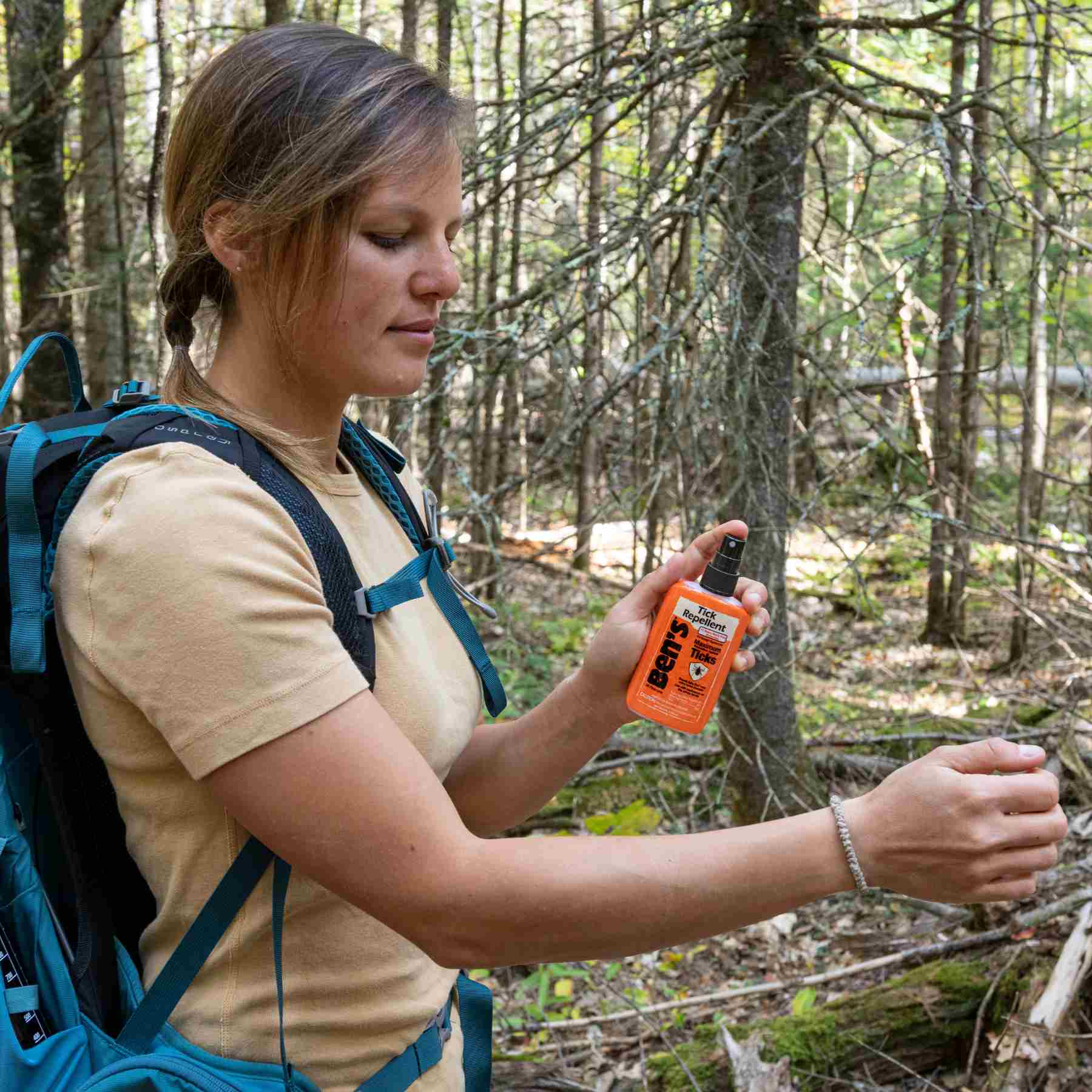 Woman using Ben's 3.4 oz Tick Repellent in woods with yellow shirt and blue backpack